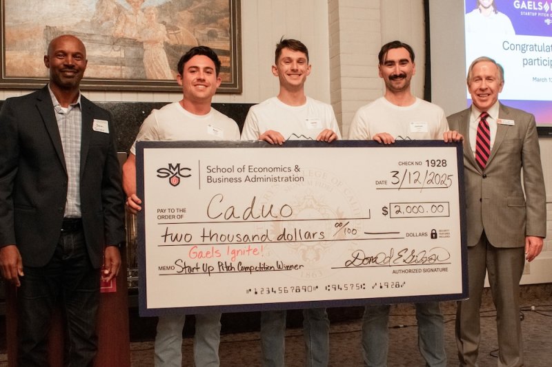 Three students representing Caduo hold a large check, with professor and dean on either side