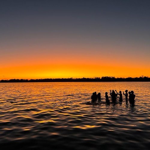 Students stand in the ocean against a bright orange sunset.