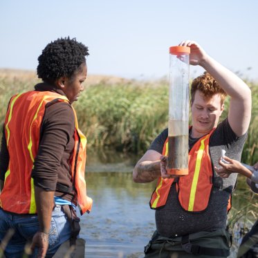 Two students are outside wearing reflective vests are gathering and observing water samples