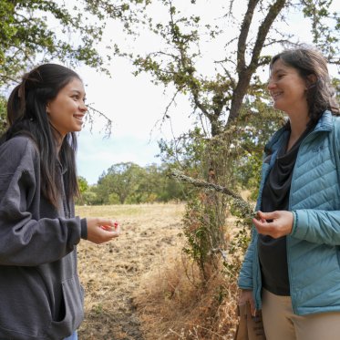 A student and professor, who is holding a tree branch, are smiling and looking at each other while outside