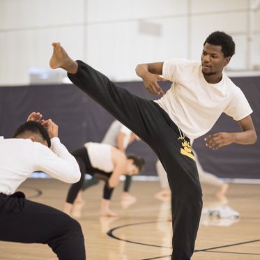 A capoeira student kicks his outstretched leg over another crouching student.