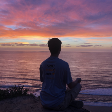 Student meditating by the ocean at sunset