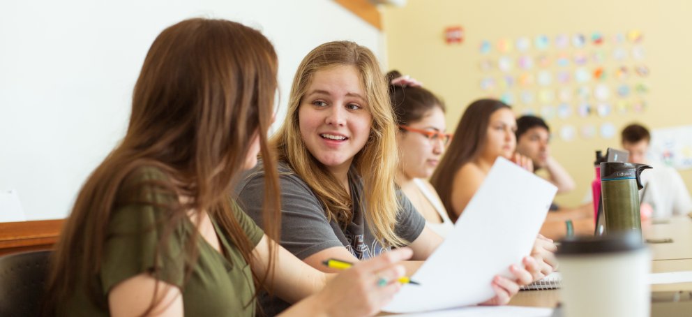 Two students in class are talking to each other while holding paper and pens