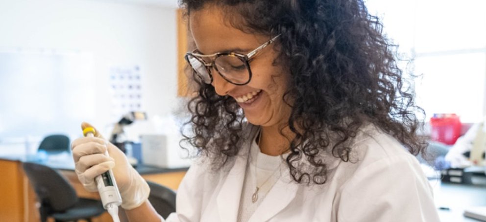 In a lab with others, a faculty member in the middle of an experiment, stands with a big smile on her face.