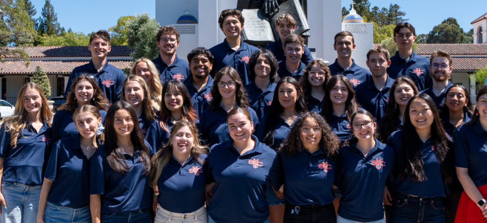 A group of resident advisors poses outdoors in front of a campus building with red-tiled roofs and a statue in the background. They are all wearing matching navy blue polo shirts with a red and white SMC logo. The group stands closely together, smiling at the camera on a sunny day.
