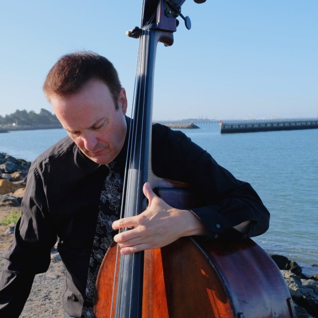 Michel Taddei, in a white dress shirt, holding a double bass. He looks down and sits with his back against the Bay