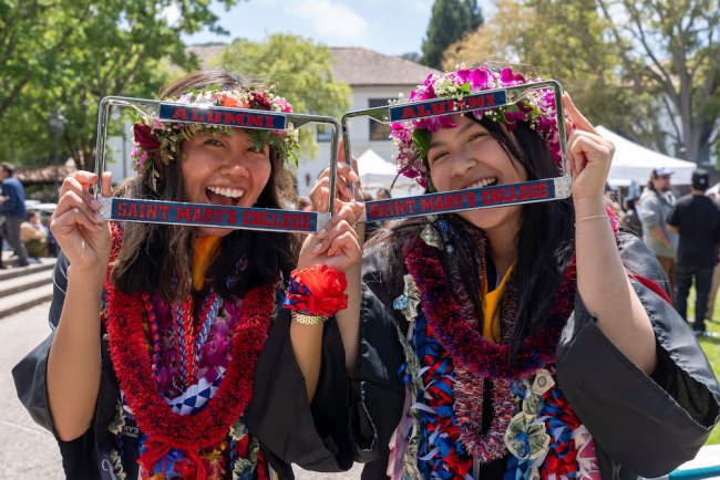 Two female students smile through Saint Mary's Alumni license plate holders after 2024 Undergraduate Commencement