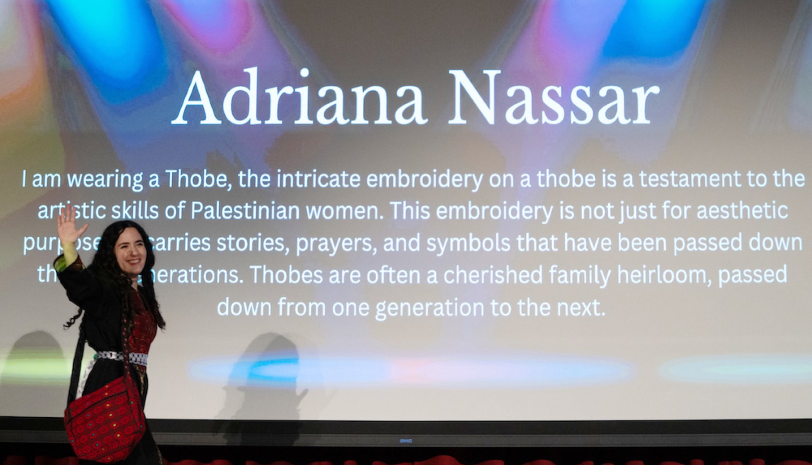 Student Adriana Nasar walks onstage in front of text describing her Thobe and its embroidery