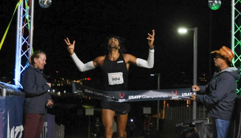 Rajpaul Pannu runs across a finish line with his arms up in the air, head looking up at the night sky