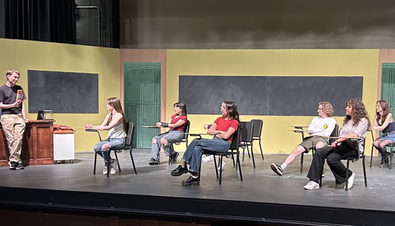 A teacher is holding up a book which the students who are seated at desks in a classroom are looking at. You can tell the class is on a stage as part of a play.