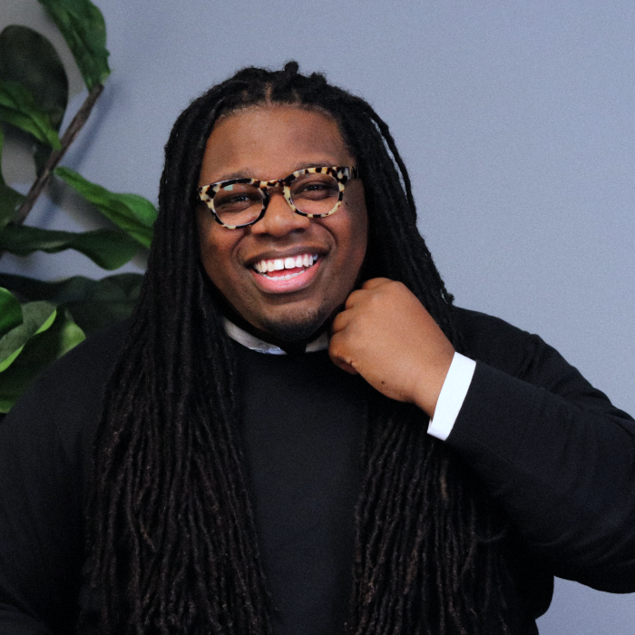 Jaron M. LeGrair smiling at camera. He is wearing black and glasses, and stands behind a hazy blue background