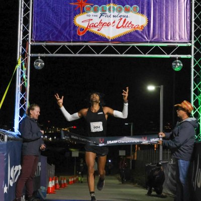 Rajpaul Pannu runs across a finish line with his arms up in the air, head looking up at the night sky