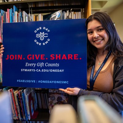 a smiling student with long brown hair holding a One Day One SMC sign