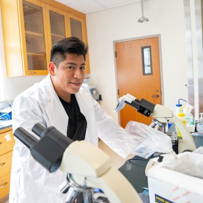male student standing in a lab coat over microscopes within a lab classroom surrounded by lab equipment, smiling at the camera