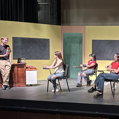 A teacher is holding up a book which the students who are seated at desks in a classroom are looking at. You can tell the class is on a stage as part of a play.