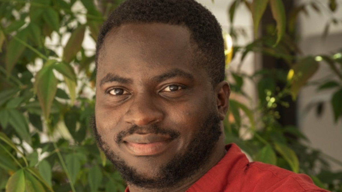 Kehinde Badiru smiles wearing a red shirt in front of trees