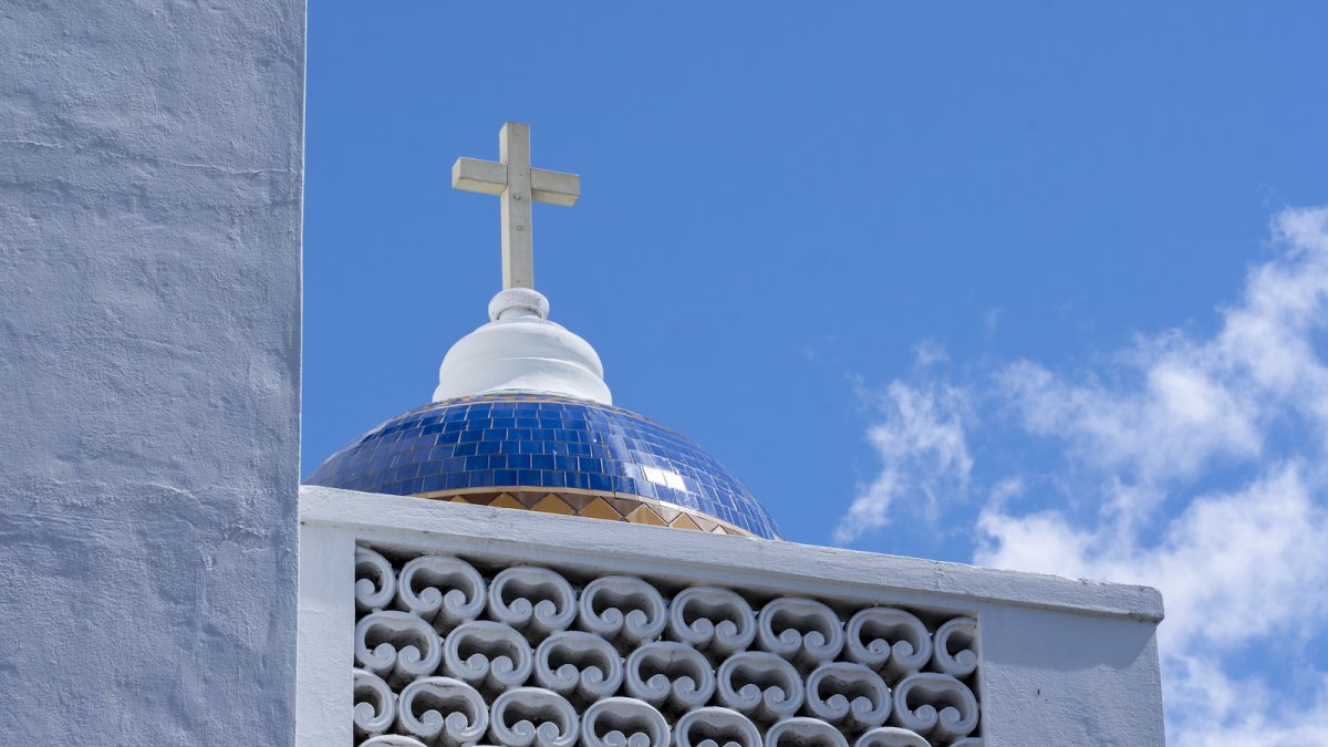 A small dome with blue tile and a white cross on top against a partly cloudy sky