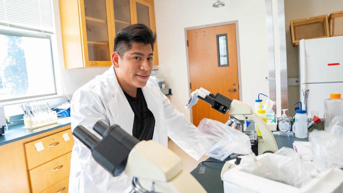 male student standing in a lab coat over microscopes within a lab classroom surrounded by lab equipment, smiling at the camera