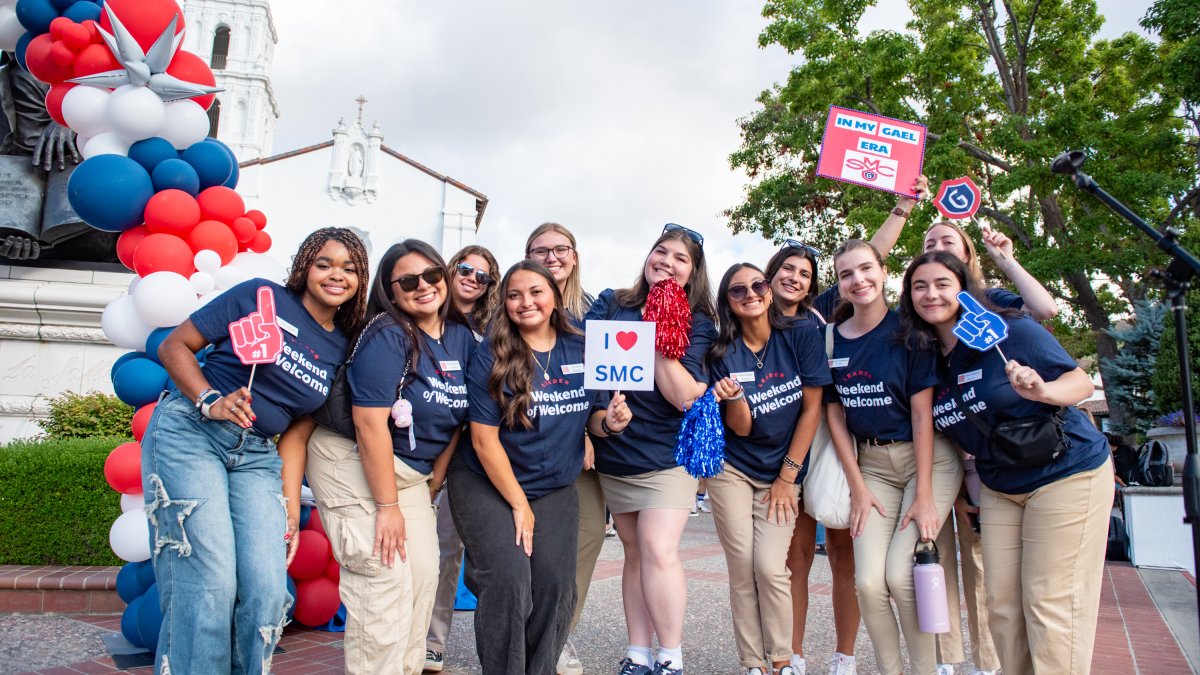 Picture of weekend of welcome leaders in front of DLS statue, posing and holding signs that say I love SMC and In My Gael Era